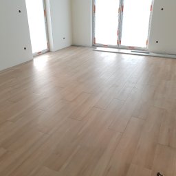 Finezja Wnętrz - Układanie paneli podłogowych w jasnym, pustym pokoju z dużymi oknami; widoczne listwy przypodłogowe i fragment ubrania w prawym dolnym rogu.