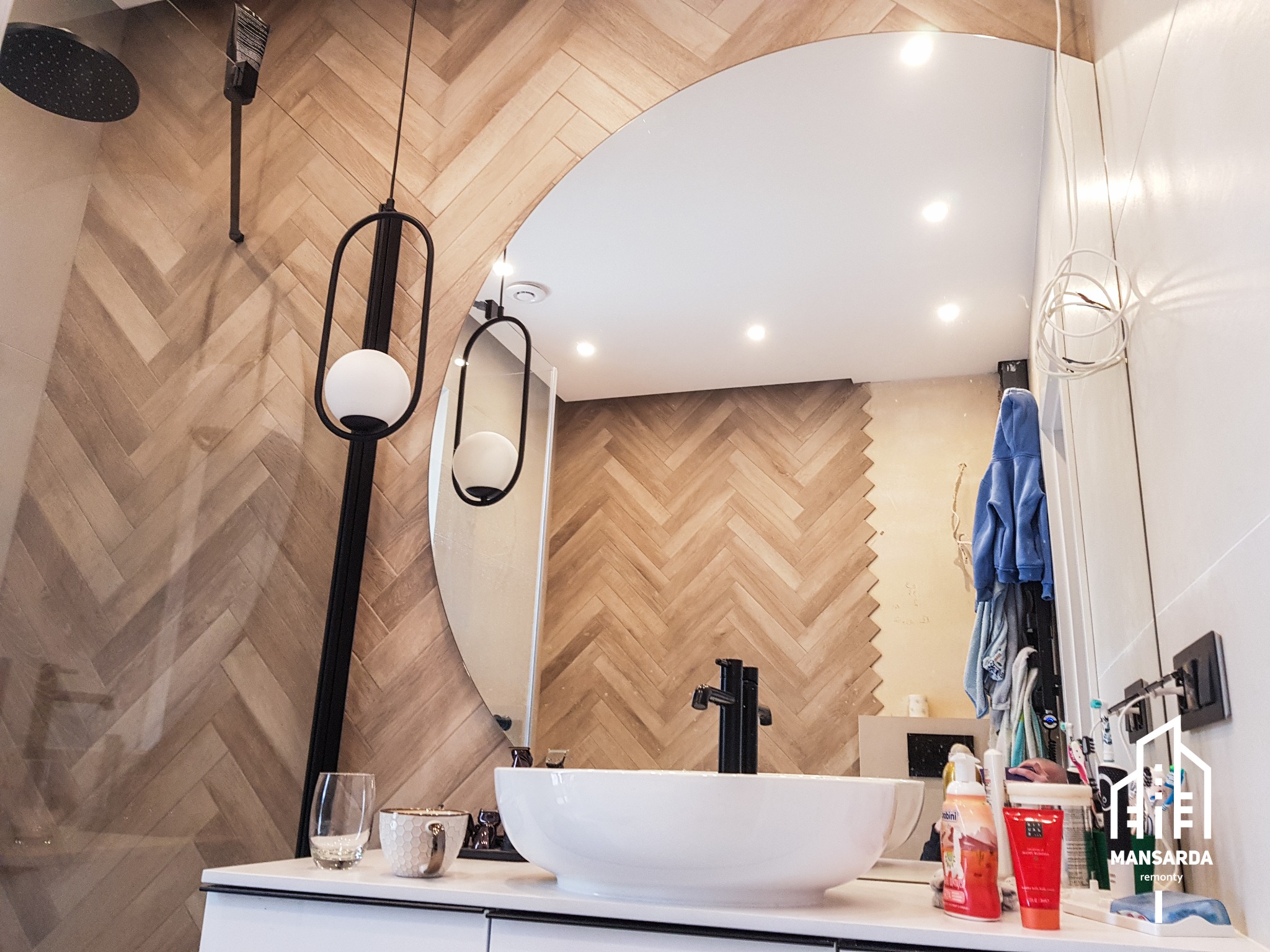 Nowoczesna łazienka z umywalką nablatową, lustrem w kształcie półkola i płytkami ułożonymi w jodełkę na ścianie, oświetlona designerską lampą wiszącą.