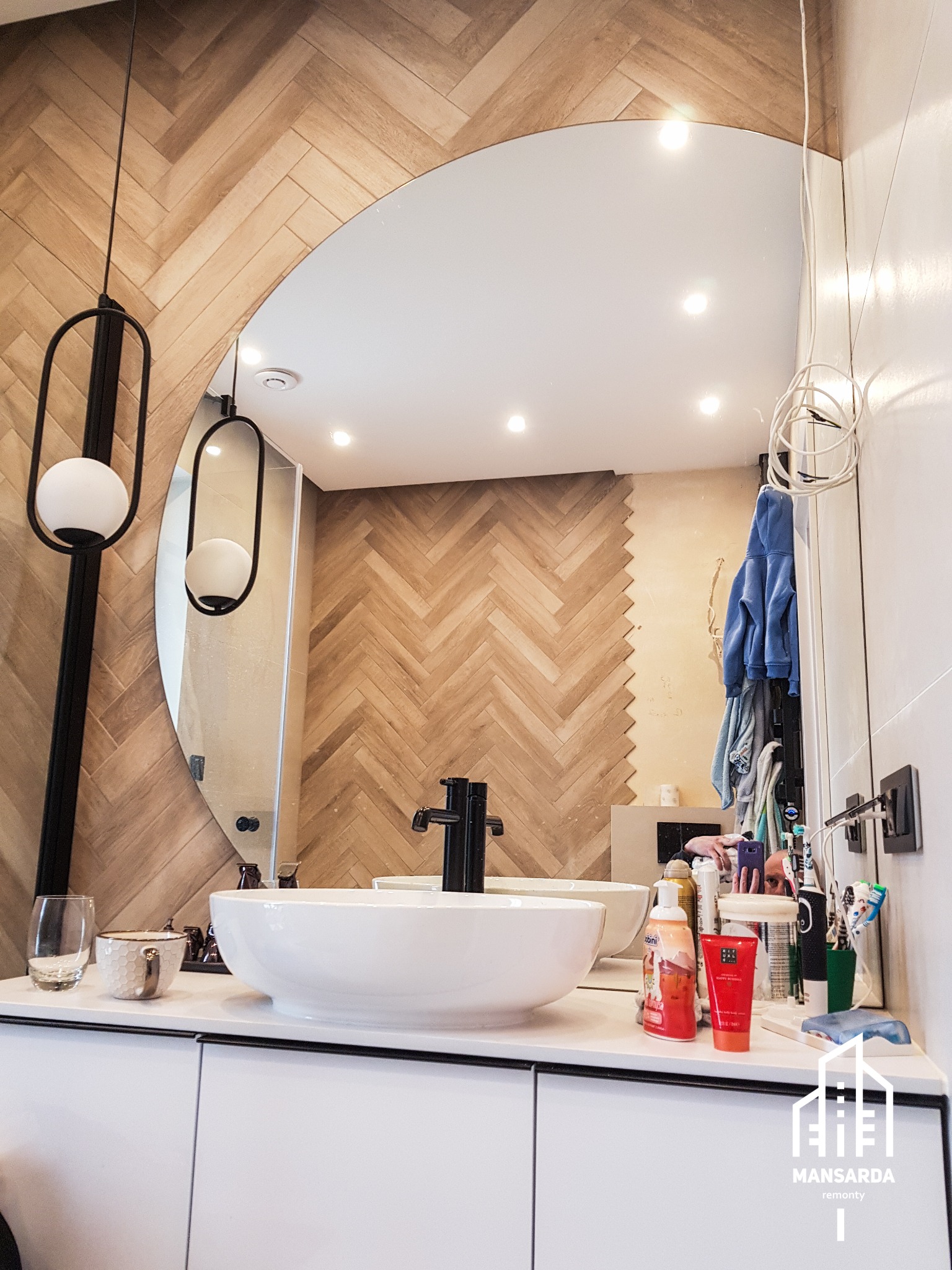 Nowoczesna łazienka z umywalką nablatową, dużym okrągłym lustrem i ścianą wykończoną płytkami imitującymi drewniany parkiet ułożony w jodełkę; widoczne wiszące lampy z okrągłymi kloszami i logo...