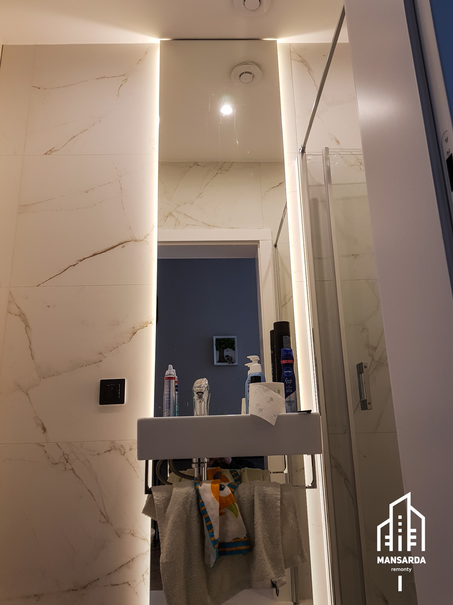 Nowoczesna łazienka z białymi płytkami imitującymi marmur, podświetlanym lustrem, minimalistyczną umywalką i kabiną prysznicową ze szklanymi drzwiami, widok z dołu na górę.