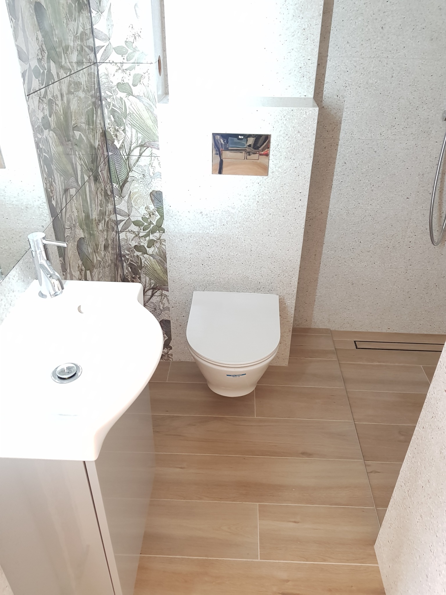 Nowoczesna łazienka z toaletą podwieszaną, umywalką nablatową na szafce, drewnopodobną podłogą i ścianą z motywem roślinnym.