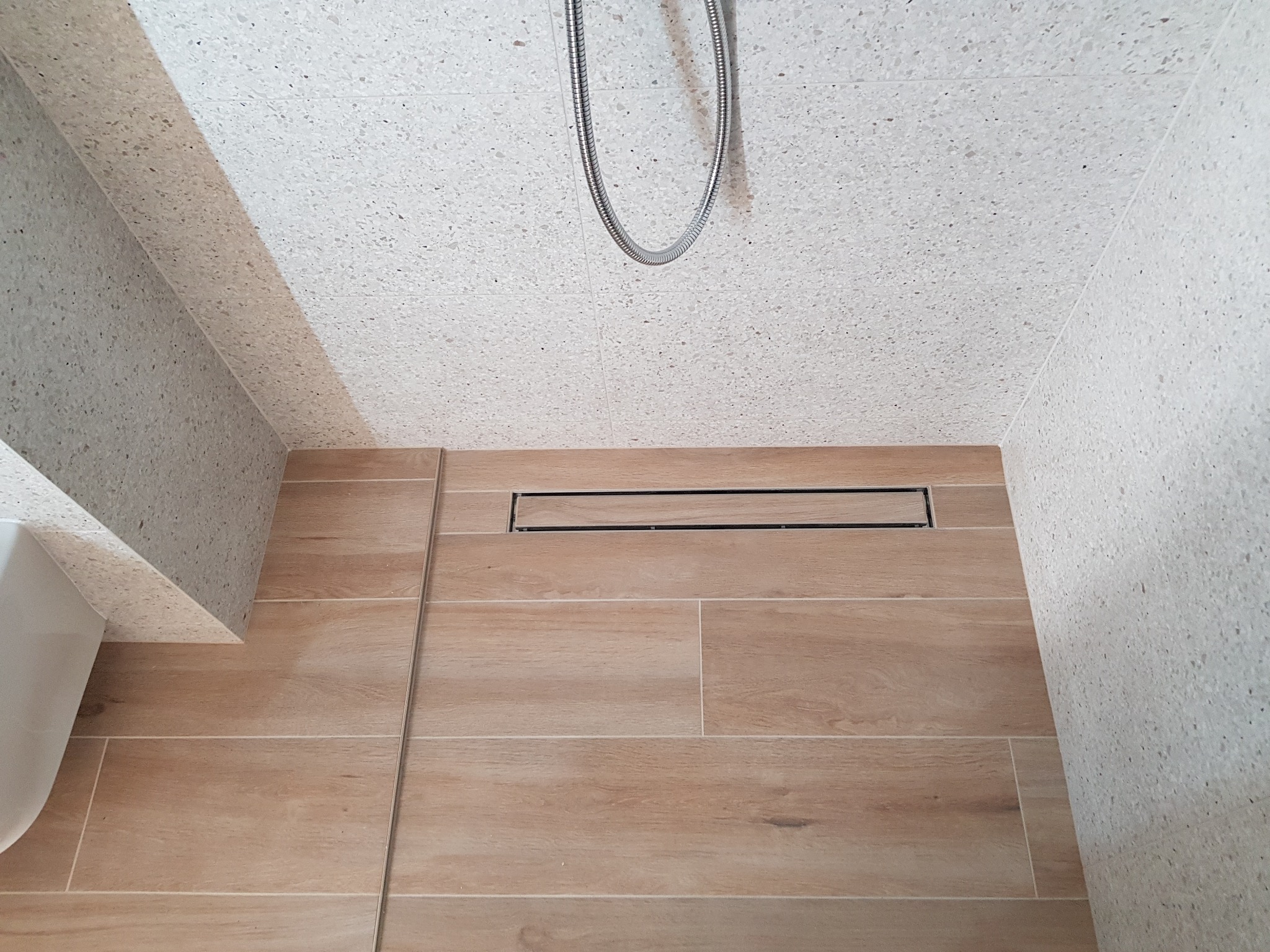 Nowoczesny odpływ liniowy w kabinie prysznicowej z beżowymi płytkami imitującymi drewno na podłodze i ścianami wykończonymi jasnym lastryko, widoczny fragment węża prysznicowego.