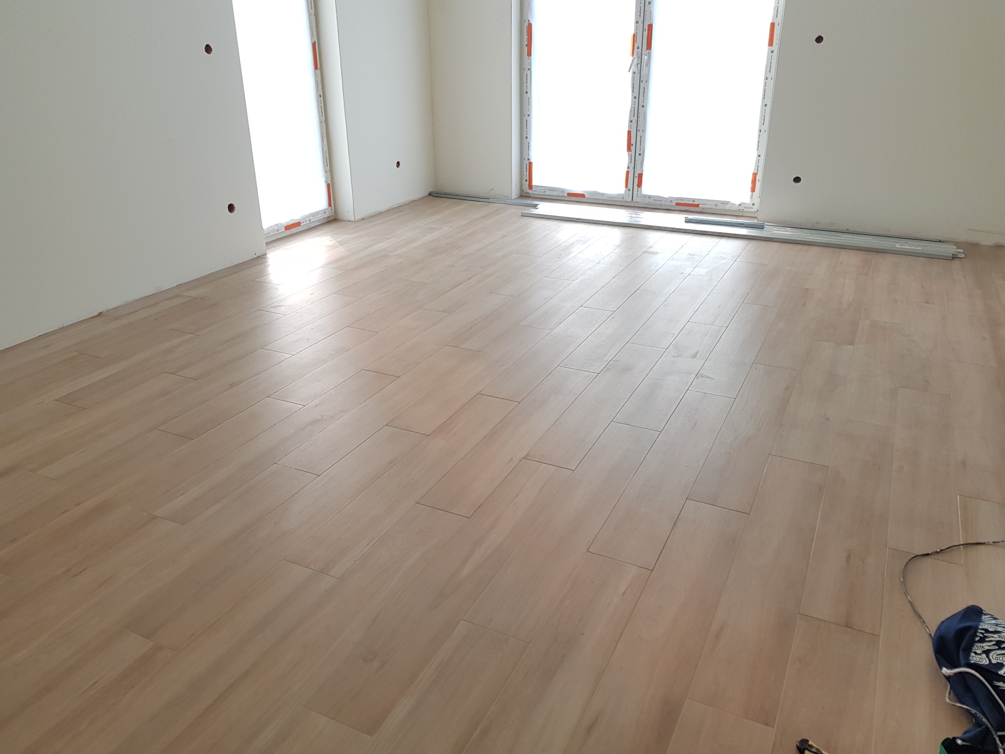 Układanie paneli podłogowych w jasnym, pustym pokoju z dużymi oknami; widoczne listwy przypodłogowe i fragment ubrania w prawym dolnym rogu.