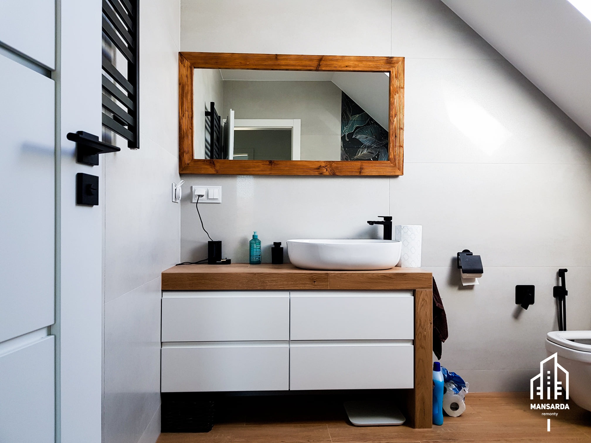 Nowoczesna łazienka na poddaszu z drewnianą ramą lustra, białą umywalką nablatową i szafką z białymi frontami oraz drewnianym blatem.