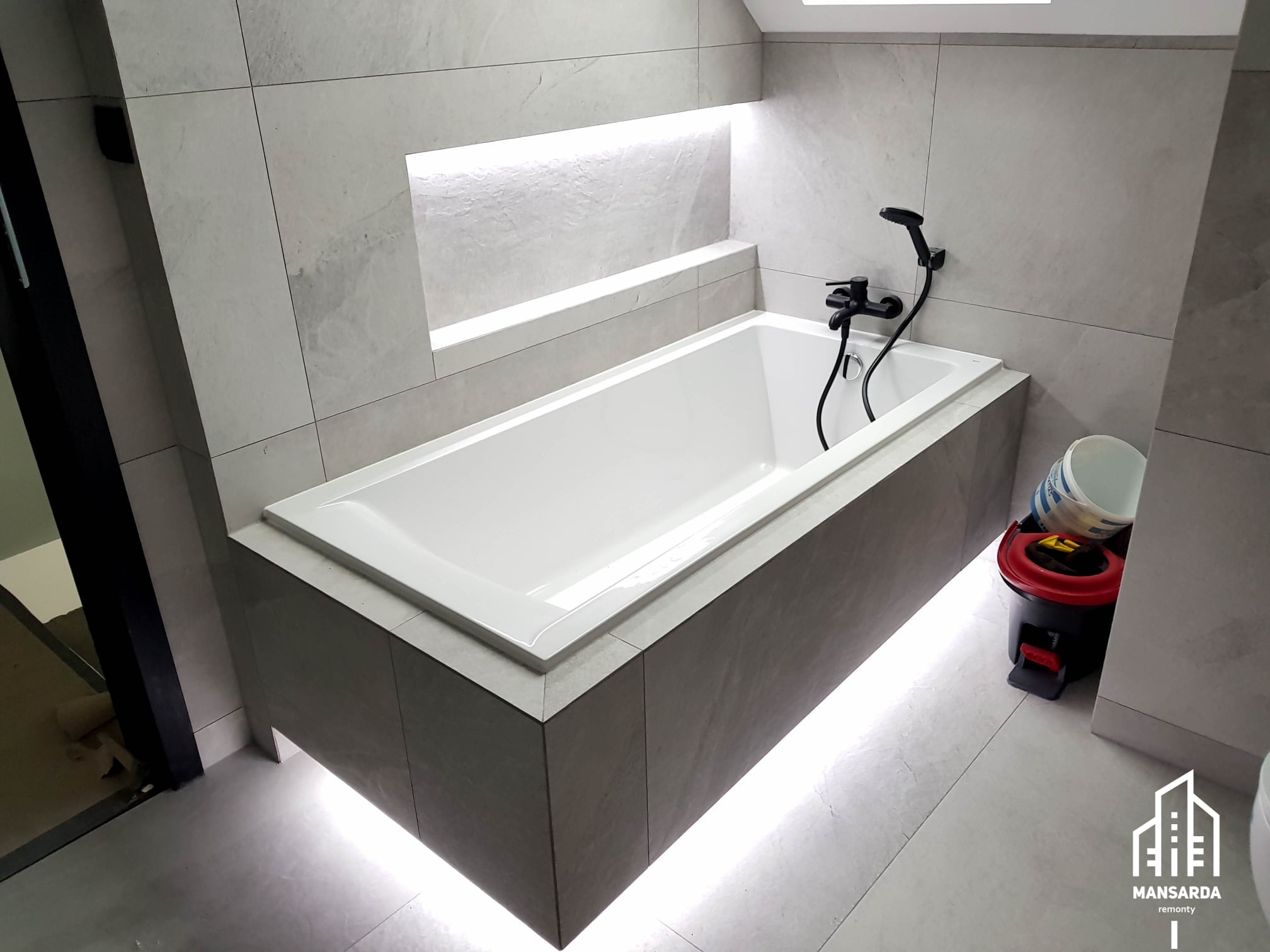 Nowoczesna łazienka w odcieniach szarości z białą wanną, podświetleniem LED pod wanną i w niszy ściennej, czarna armatura i widoczne narzędzia malarskie.