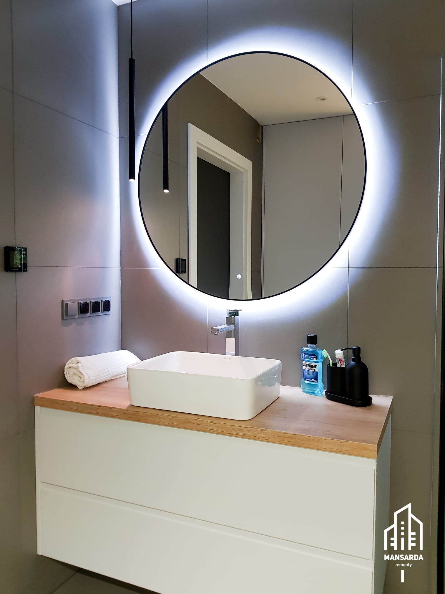 Nowoczesna łazienka z okrągłym lustrem LED, białą umywalką nablatową na drewnianym blacie i szarą płytką ścienną. Minimalistyczne oświetlenie punktowe i logo firmy w prawym dolnym rogu.