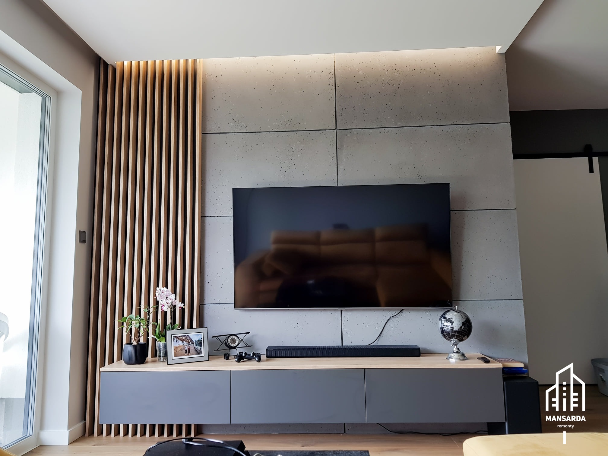 Nowoczesny salon z szarą ścianą wykończoną betonem architektonicznym, drewnianymi lamelami i szafką RTV w kolorze szarym z drewnianym blatem, oświetlenie LED pod sufitem.