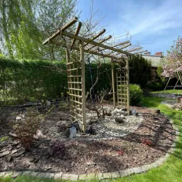 Drewniana konstrukcja z kratownicami, prawdopodobnie pod pnącza, otoczona kamieniami i korą na trawniku w ogrodzie, w tle zieleń drzew i krzewów.