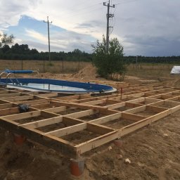Daniel Fryc Drewniany Danny - Konstrukcja drewniana tarasu w trakcie budowy, widoczny szkielet z belek na betonowych podporach, obok basen ogrodowy, w tle drzewa i słupy elektryczne.