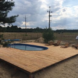 Daniel Fryc Drewniany Danny - Drewniany podest z basenem wkomponowany w piaszczysty teren, z drewnianymi leżakami i stolikiem, w tle łąka i słupy energetyczne.