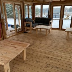 Daniel Fryc Drewniany Danny - Wnętrze przeszklonej konstrukcji z drewnianą podłogą i prostymi meblami z jasnego drewna, widok na zaśnieżony krajobraz przez okna.