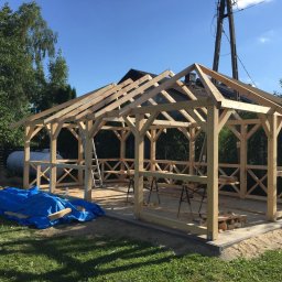 Daniel Fryc Drewniany Danny - Konstrukcja drewniana altany w trakcie budowy, widoczne elementy więźby dachowej i barierki z krzyżowymi wzorami, niebieska plandeka zakrywa materiały budowlane, w tle zbiornik gazu.