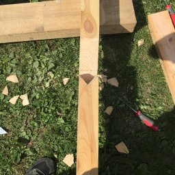 Daniel Fryc Drewniany Danny - Przygotowanie elementu drewnianego z wyciętym gniazdem w kształcie litery V na trawie, widoczne narzędzia i fragment buta.