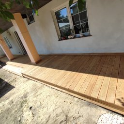 Daniel Fryc Drewniany Danny - Nowy, jasny taras z drewna, z widocznymi słupami podtrzymującymi zadaszenie, przylegający do białego budynku z oknem, na tle piaszczystego podłoża.