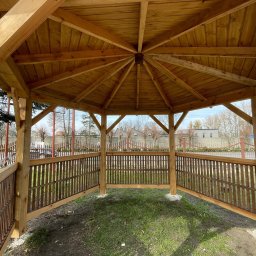 Daniel Fryc Drewniany Danny - Drewniana konstrukcja zadaszona, widok od wewnątrz, z brązowymi balustradami, na trawiastym podłożu, w tle zabudowa jednorodzinna.