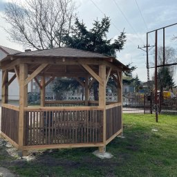 Daniel Fryc Drewniany Danny - Nowo wybudowana, drewniana altana z brązowym dachem i panelami w dolnej części, umieszczona na trawniku przed budynkiem mieszkalnym.