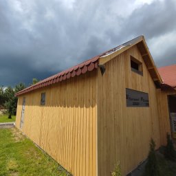 Daniel Fryc Drewniany Danny - Budynek w konstrukcji szkieletowej z drewnianą elewacją pionową i dachem pokrytym gontem bitumicznym w kształcie rybiej łuski, widoczne membrany paroizolacyjne na oknach, pochmurne niebo w tle.