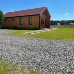 Nowoczesny, parterowy dom z drewna z czerwonym dachem, widok z zewnątrz na tle błękitnego nieba i zieleni, z żwirowym podjazdem i dwoma krzesłami na małym tarasie.