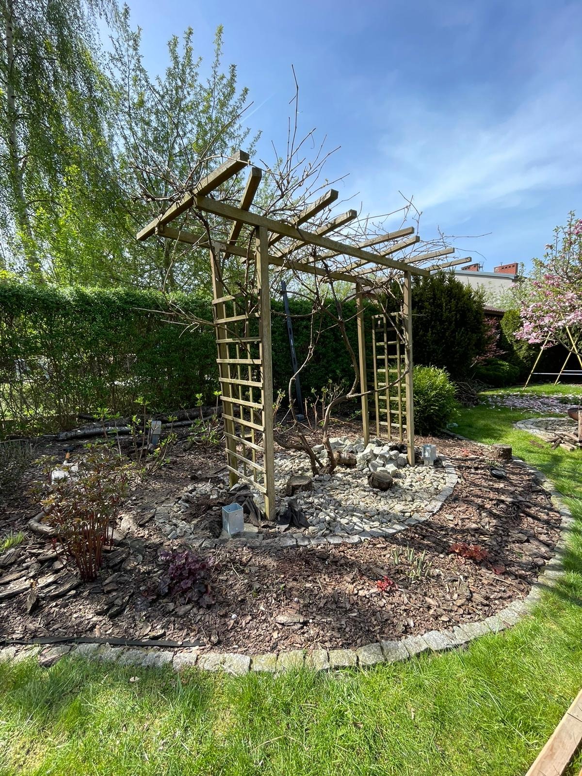 Drewniana konstrukcja z kratownicami, prawdopodobnie pod pnącza, otoczona kamieniami i korą na trawniku w ogrodzie, w tle zieleń drzew i krzewów.