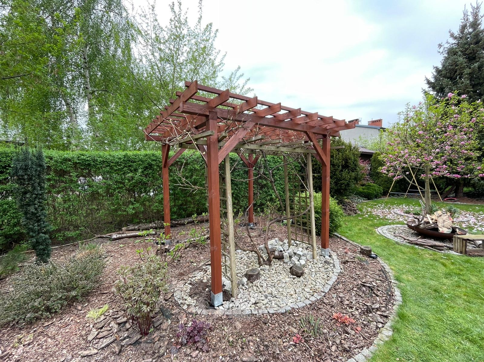 Brązowa, drewniana konstrukcja z kratownicą wewnątrz, otoczona kamieniami i korą, w tle zielony żywopłot i drzewa.
