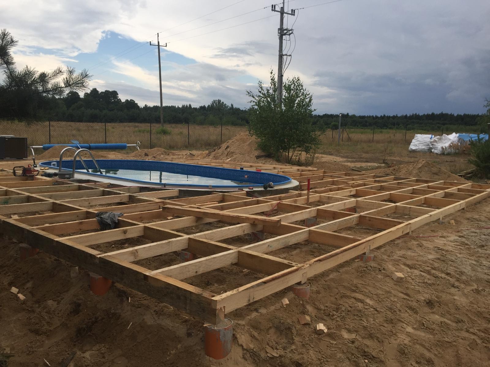 Konstrukcja drewniana tarasu w trakcie budowy, widoczny szkielet z belek na betonowych podporach, obok basen ogrodowy, w tle drzewa i słupy elektryczne.