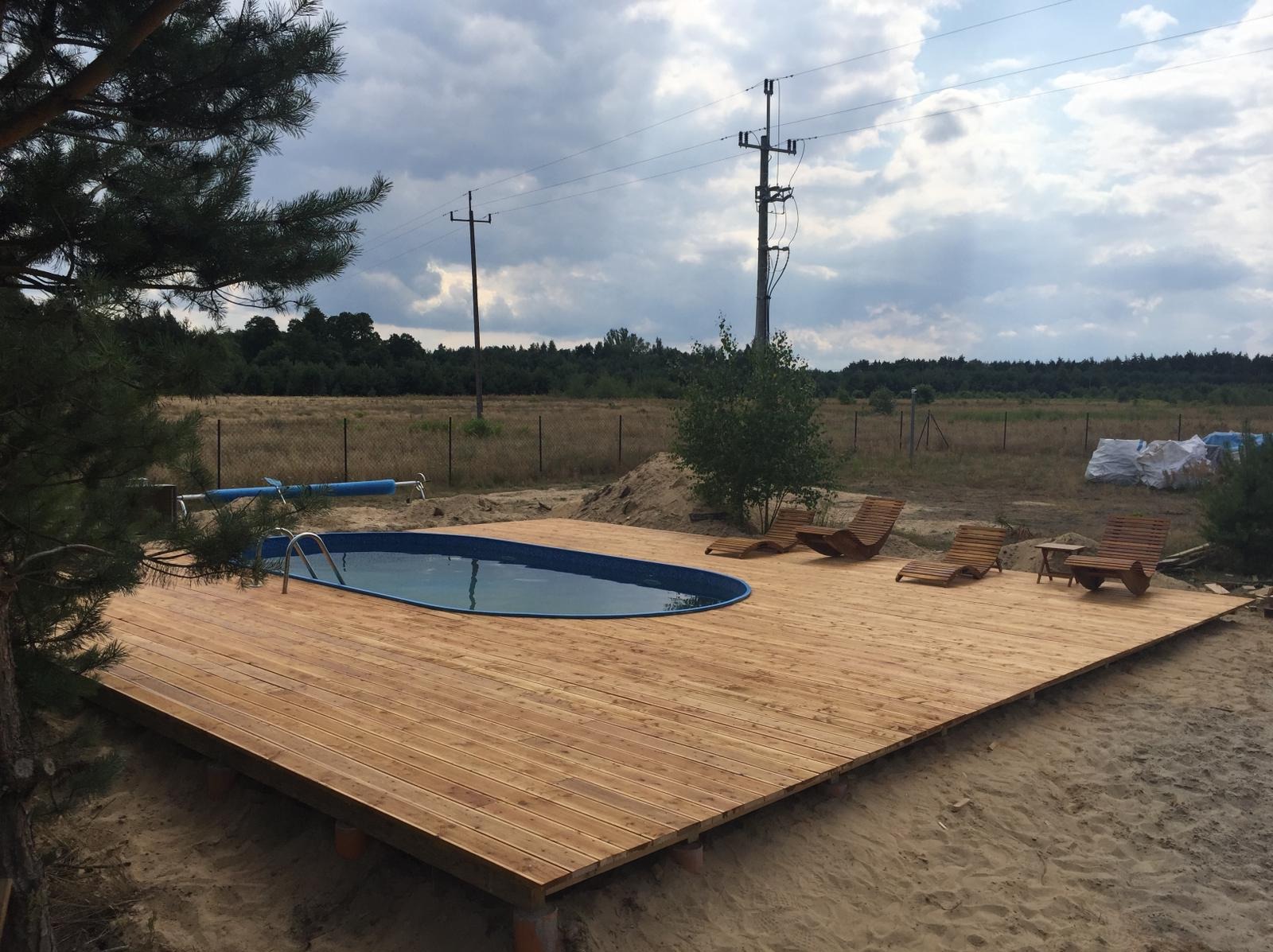Drewniany podest z basenem wkomponowany w piaszczysty teren, z drewnianymi leżakami i stolikiem, w tle łąka i słupy energetyczne.