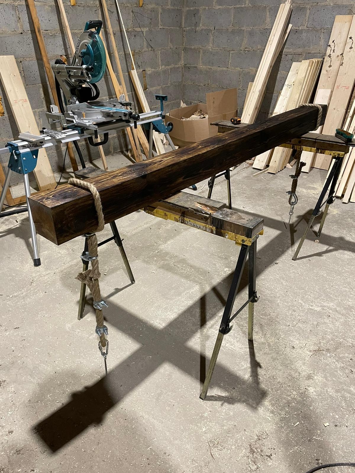 Obrabiana drewniana belka o ciemnym wybarwieniu, umieszczona na kozłach warsztatowych, z liną dekoracyjną, w tle ukośnica i deski.