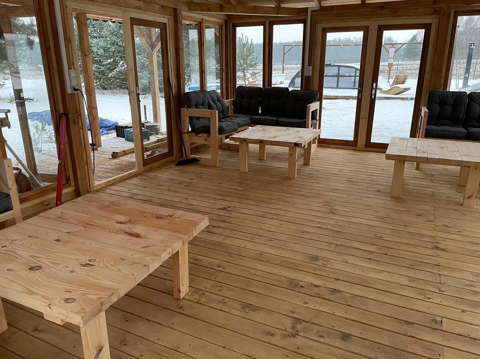 Wnętrze przeszklonej konstrukcji z drewnianą podłogą i prostymi meblami z jasnego drewna, widok na zaśnieżony krajobraz przez okna.