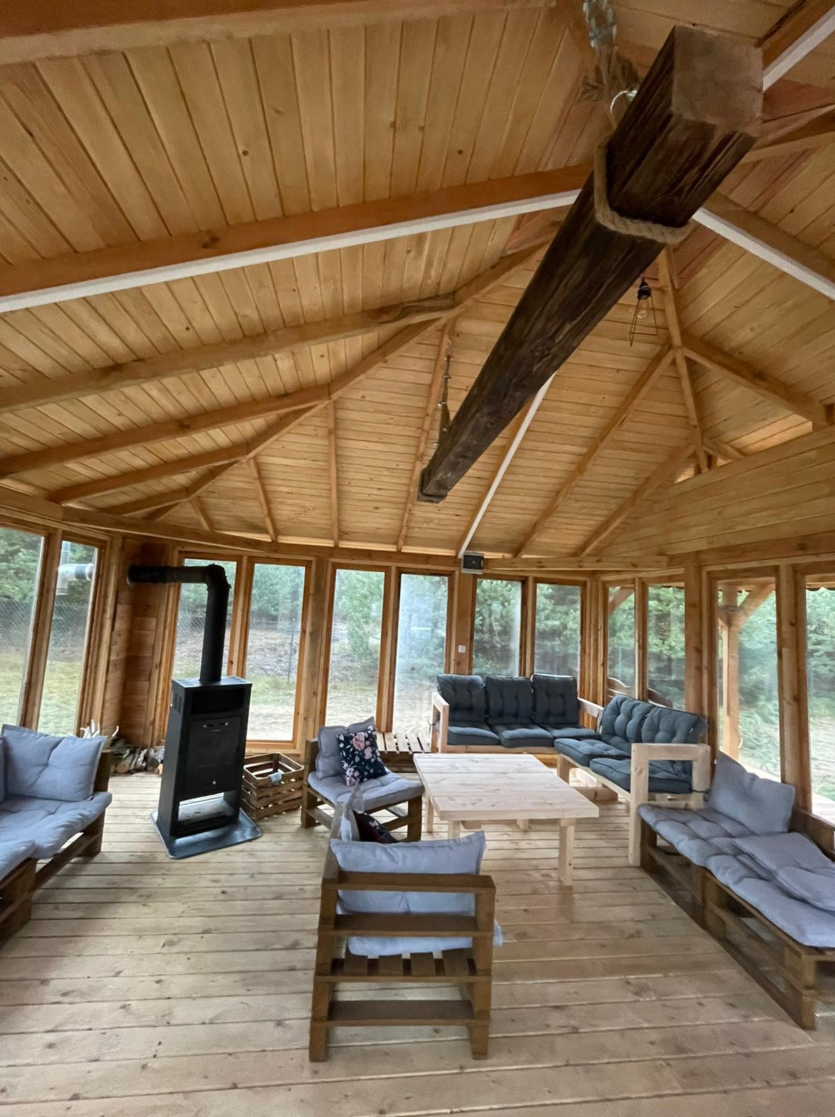 Wnętrze drewnianej konstrukcji z dużymi oknami, wyposażone w drewniane meble z szarymi poduszkami, piec typu koza i ozdobną belkę pod sufitem.