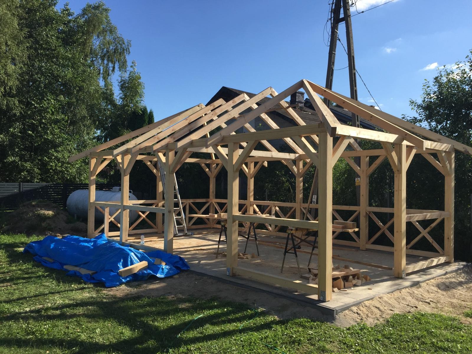 Konstrukcja drewniana altany w trakcie budowy, widoczne elementy więźby dachowej i barierki z krzyżowymi wzorami, niebieska plandeka zakrywa materiały budowlane, w tle zbiornik gazu.