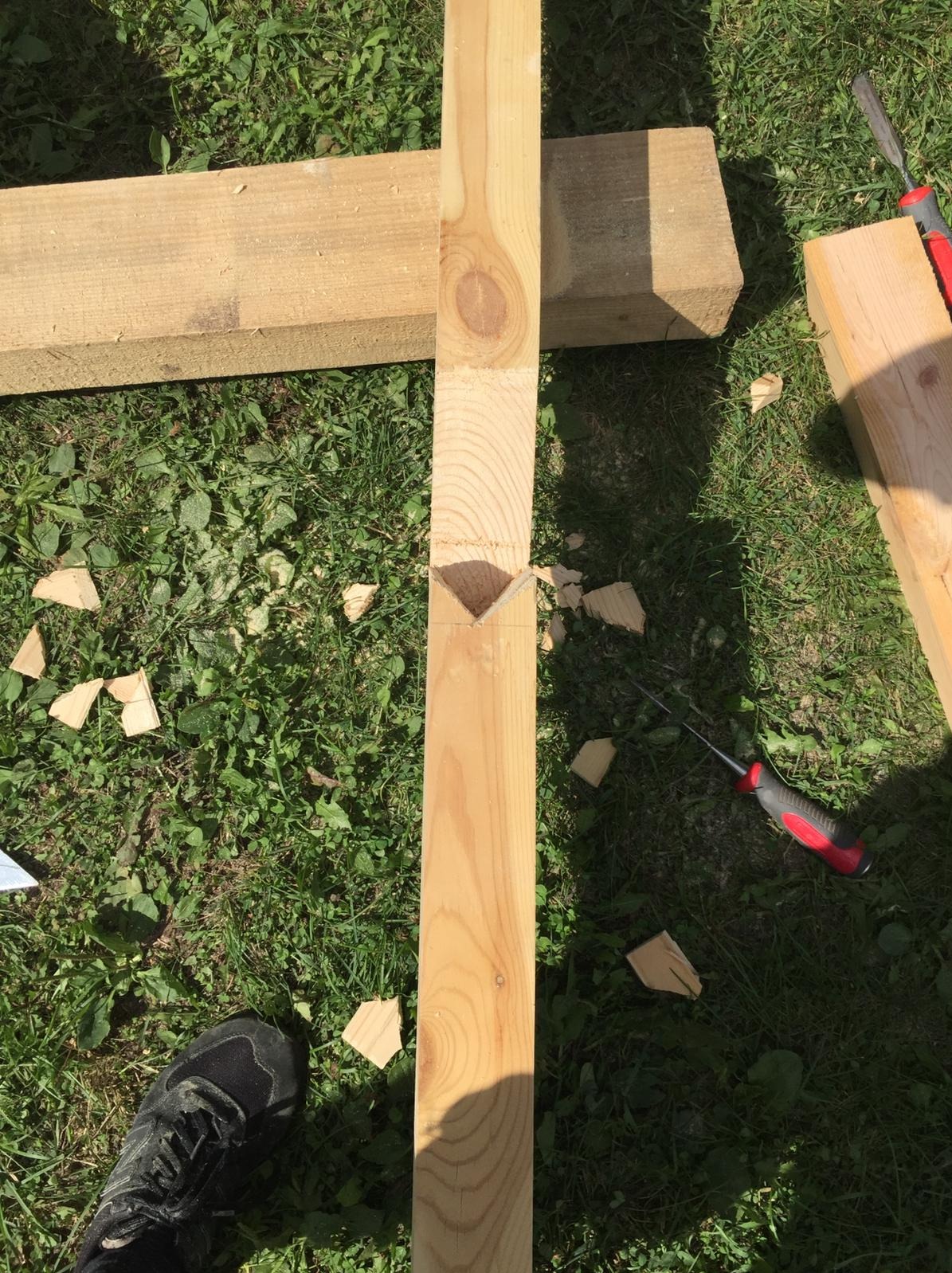 Przygotowanie elementu drewnianego z wyciętym gniazdem w kształcie litery V na trawie, widoczne narzędzia i fragment buta.