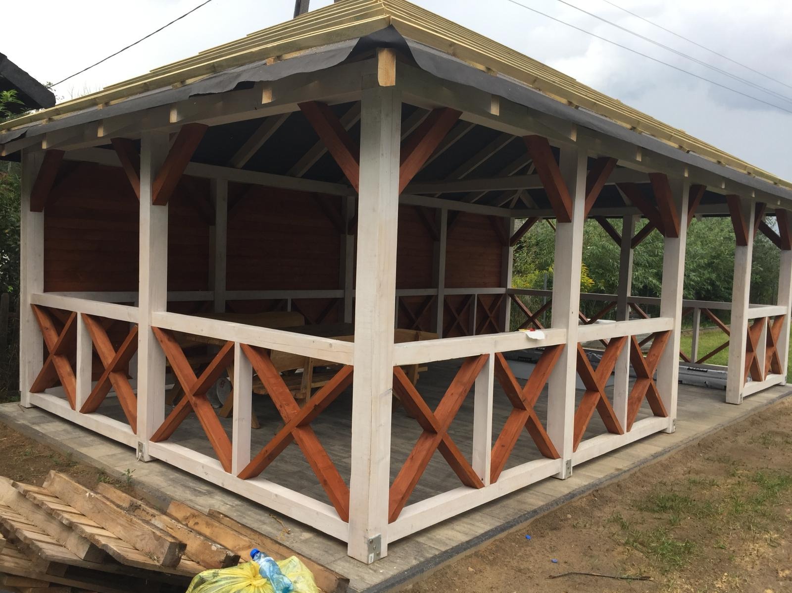 Drewniana altana z białymi słupami i pomarańczowo-brązowymi krzyżowymi elementami dekoracyjnymi, częściowo zadaszona, na kostce brukowej, z widocznymi materiałami budowlanymi obok.