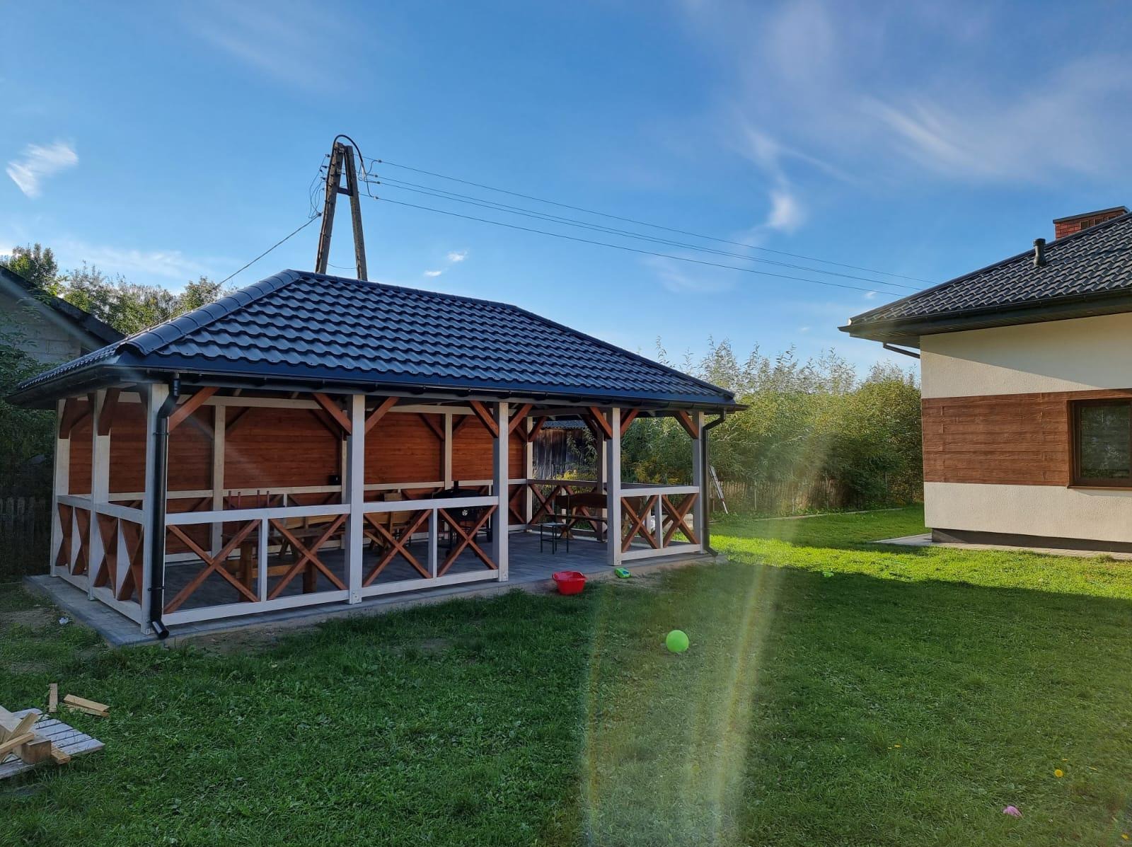 Nowoczesna altana z ciemnym dachem i jasnymi elementami drewnianymi, widoczna z zewnątrz na trawniku w słoneczny dzień, z zabawkami dziecięcymi w pobliżu.