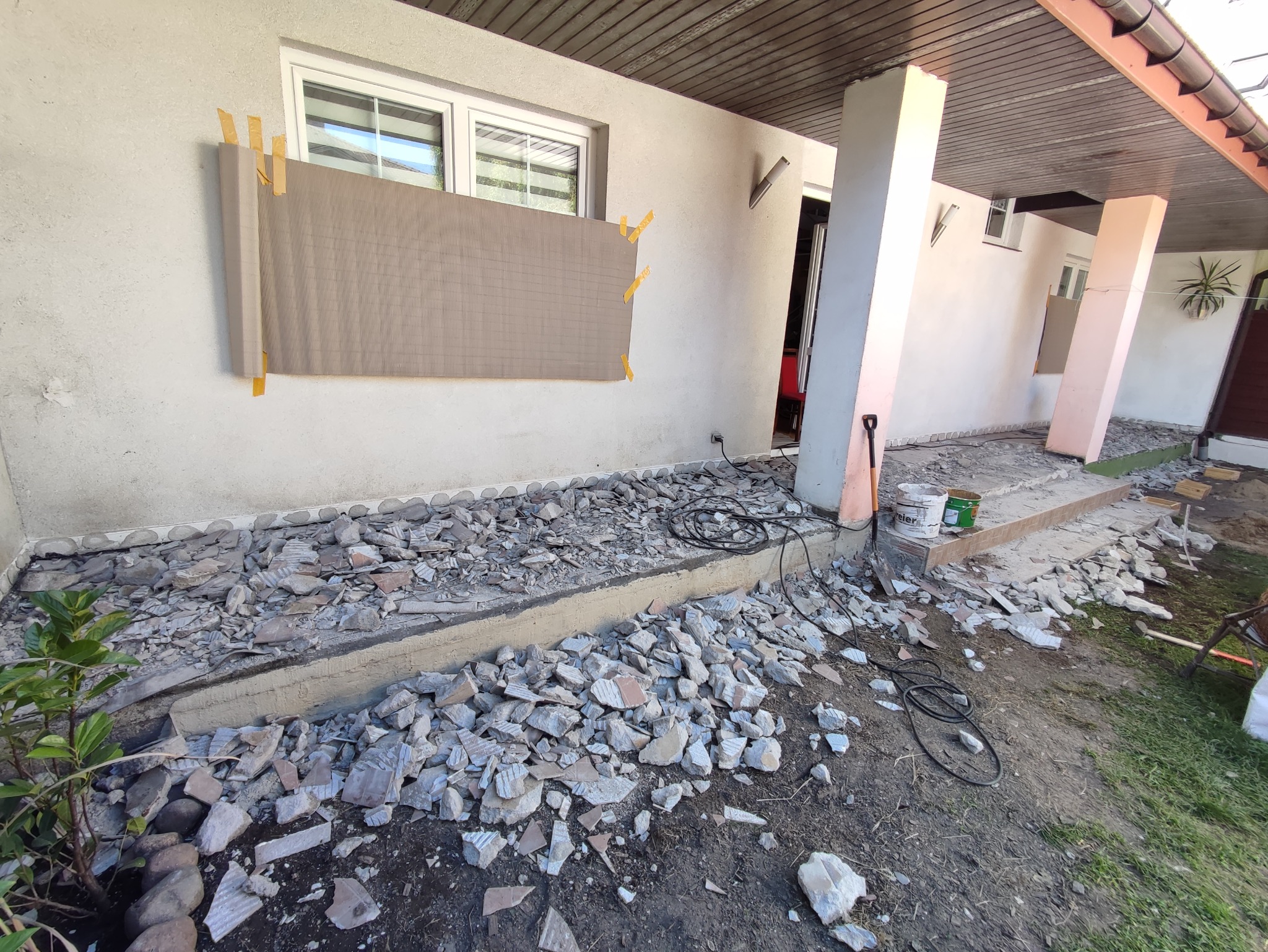 Przygotowanie elewacji domu do remontu: fragment ściany z przyklejoną płytą izolacyjną, gruz po skuciu starych płytek tarasowych, widoczne narzędzia i wiaderka z farbą.