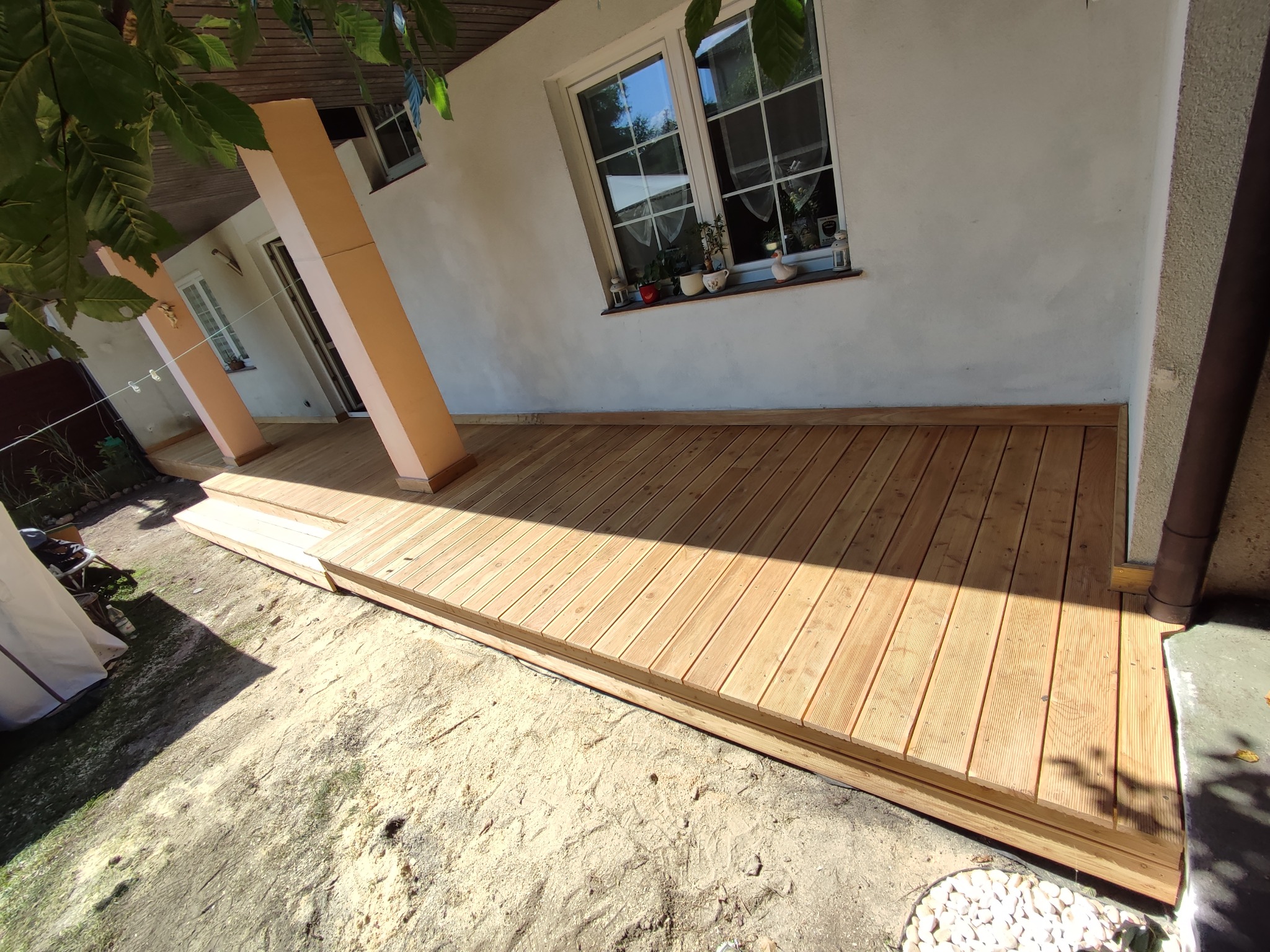 Nowy, jasny taras z drewna, z widocznymi słupami podtrzymującymi zadaszenie, przylegający do białego budynku z oknem, na tle piaszczystego podłoża.