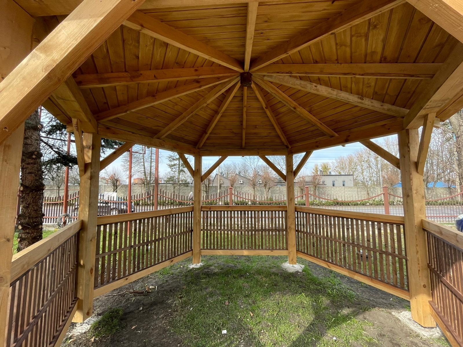 Drewniana konstrukcja zadaszona, widok od wewnątrz, z brązowymi balustradami, na trawiastym podłożu, w tle zabudowa jednorodzinna.