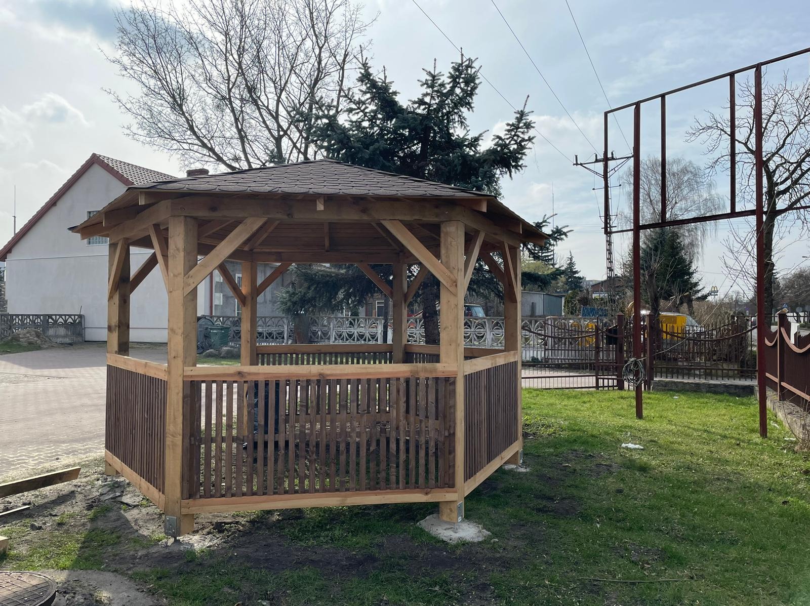 Nowo wybudowana, drewniana altana z brązowym dachem i panelami w dolnej części, umieszczona na trawniku przed budynkiem mieszkalnym.