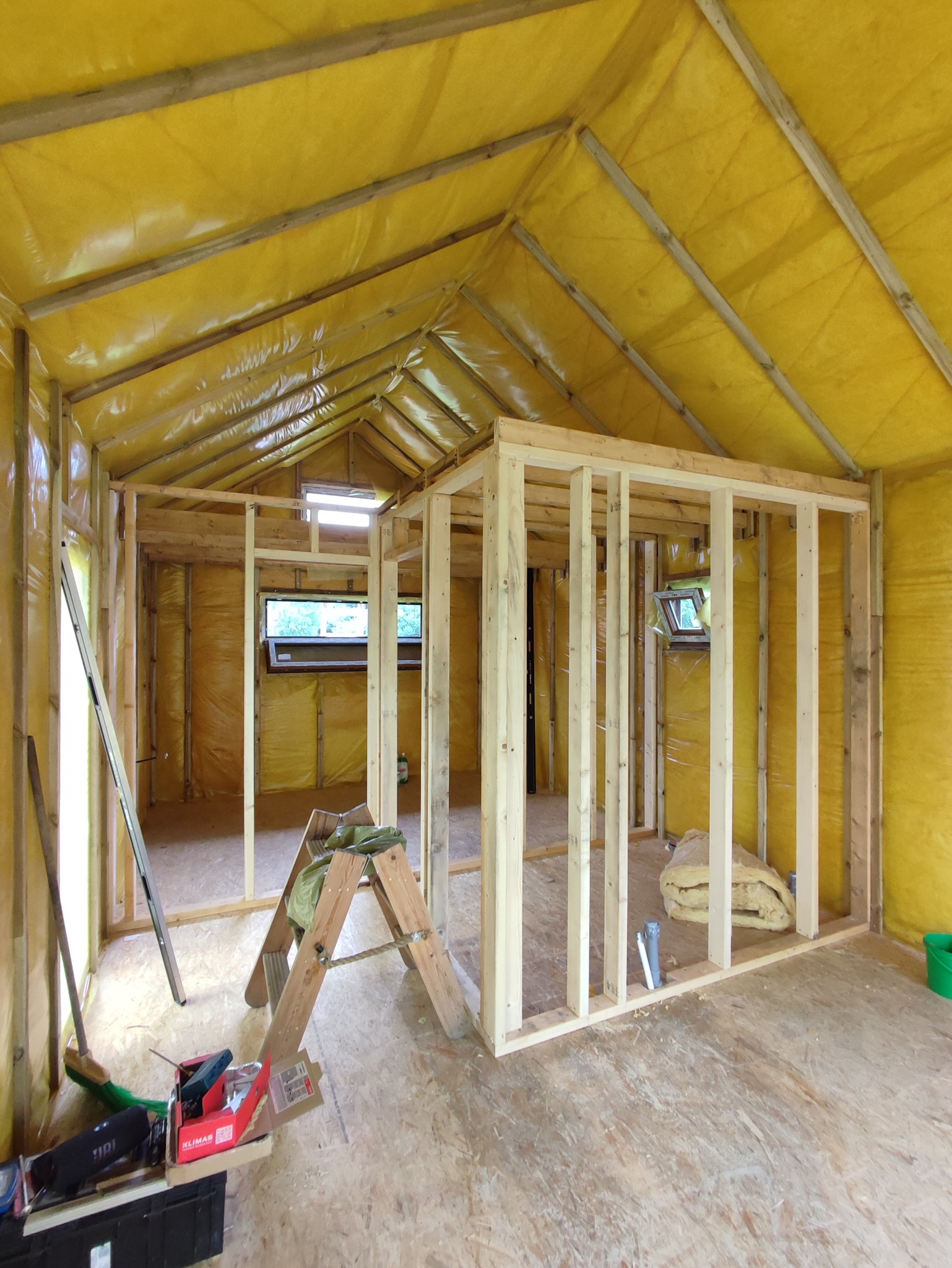 Wnętrze domu szkieletowego w trakcie budowy: widoczne drewniane ramy ścian działowych, ocieplenie ścian folią w kolorze żółtym, narzędzia i drabina.