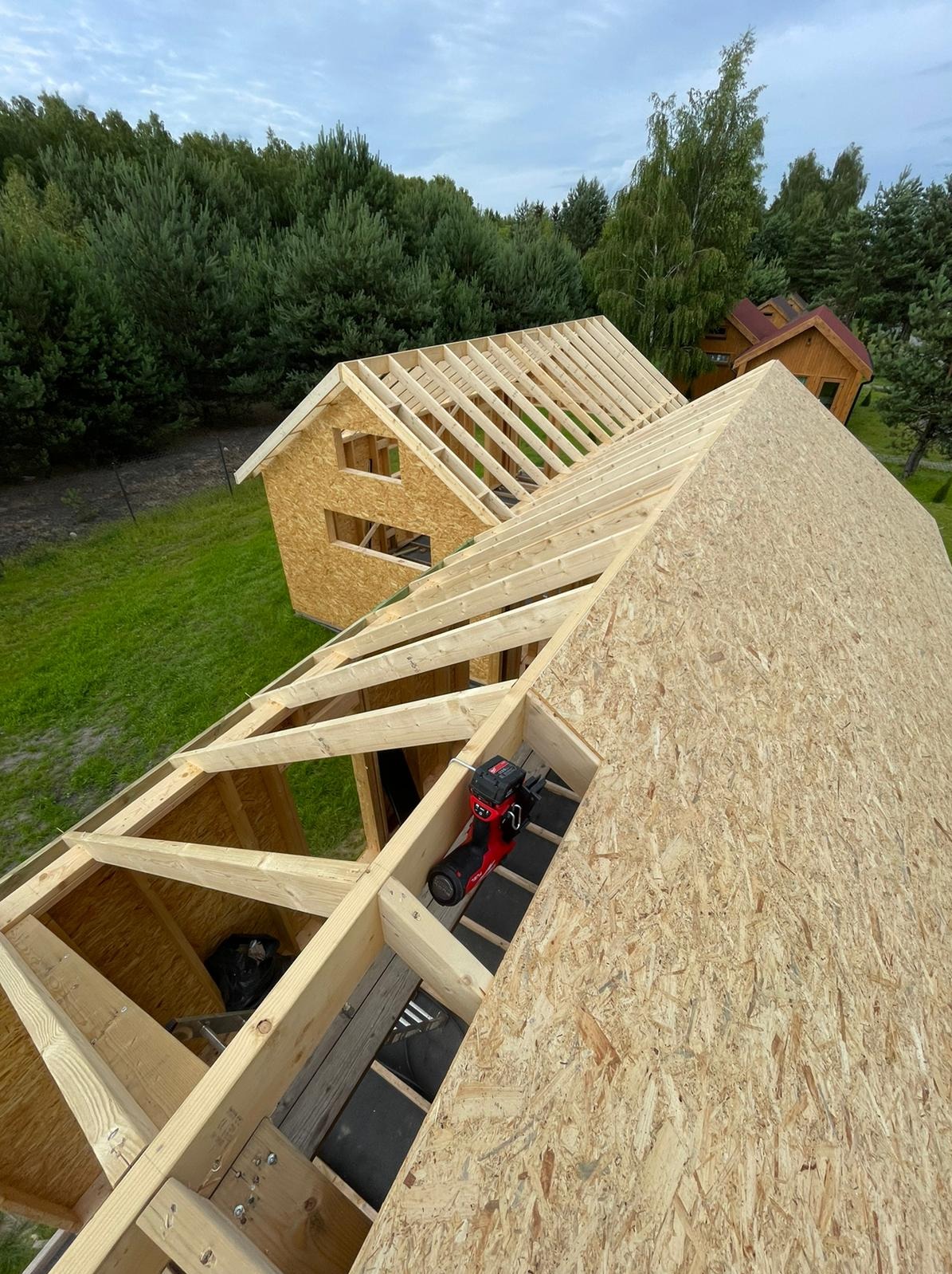 Wznoszenie szkieletu drewnianego domu z widoczną konstrukcją dachu i narzędziem do montażu na poszyciu z płyty OSB, w tle zieleń i inne budynki.