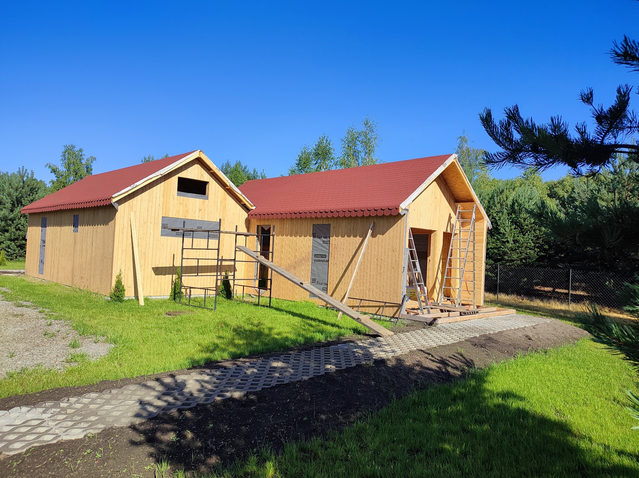 Dwa drewniane budynki w trakcie budowy z czerwonymi dachami, widoczne rusztowania i drabiny na tle zielonego trawnika i błękitnego nieba.