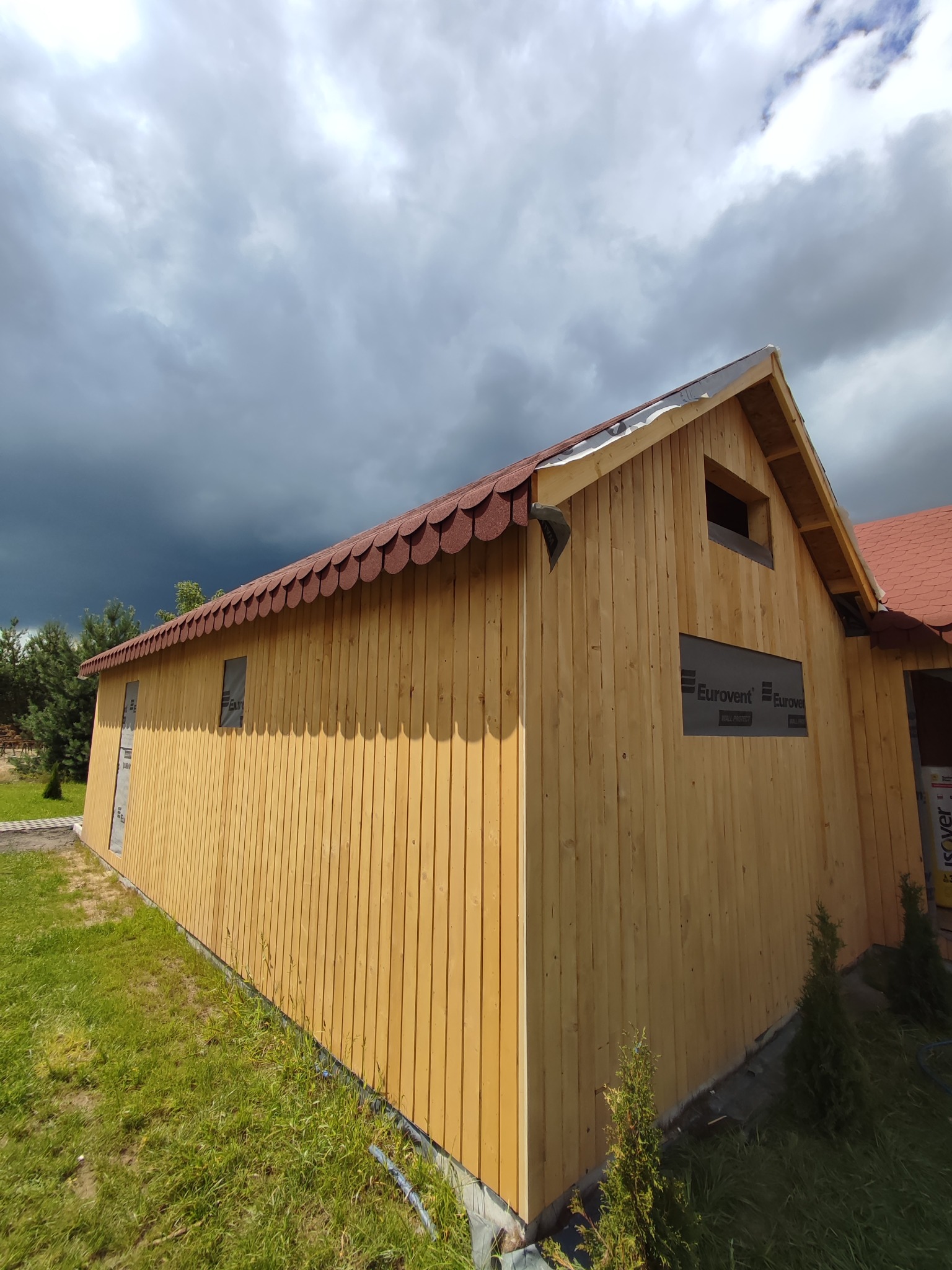 Budynek w konstrukcji szkieletowej z drewnianą elewacją pionową i dachem pokrytym gontem bitumicznym w kształcie rybiej łuski, widoczne membrany paroizolacyjne na oknach, pochmurne niebo w tle.