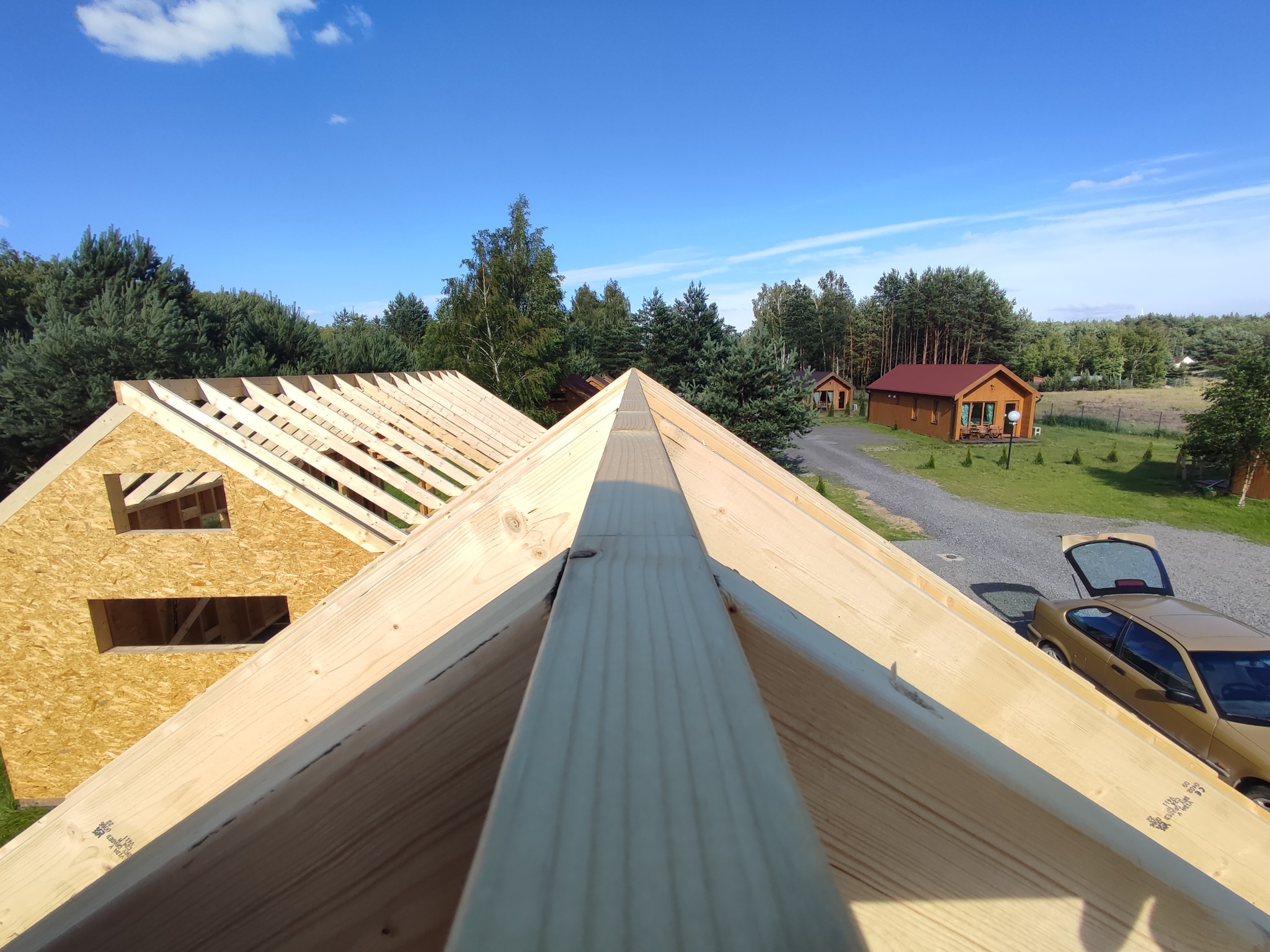 Konstrukcja dachu w trakcie budowy, widok z góry na drewniane krokwie i deskowanie, w tle domki letniskowe i samochód z otwartym bagażnikiem.