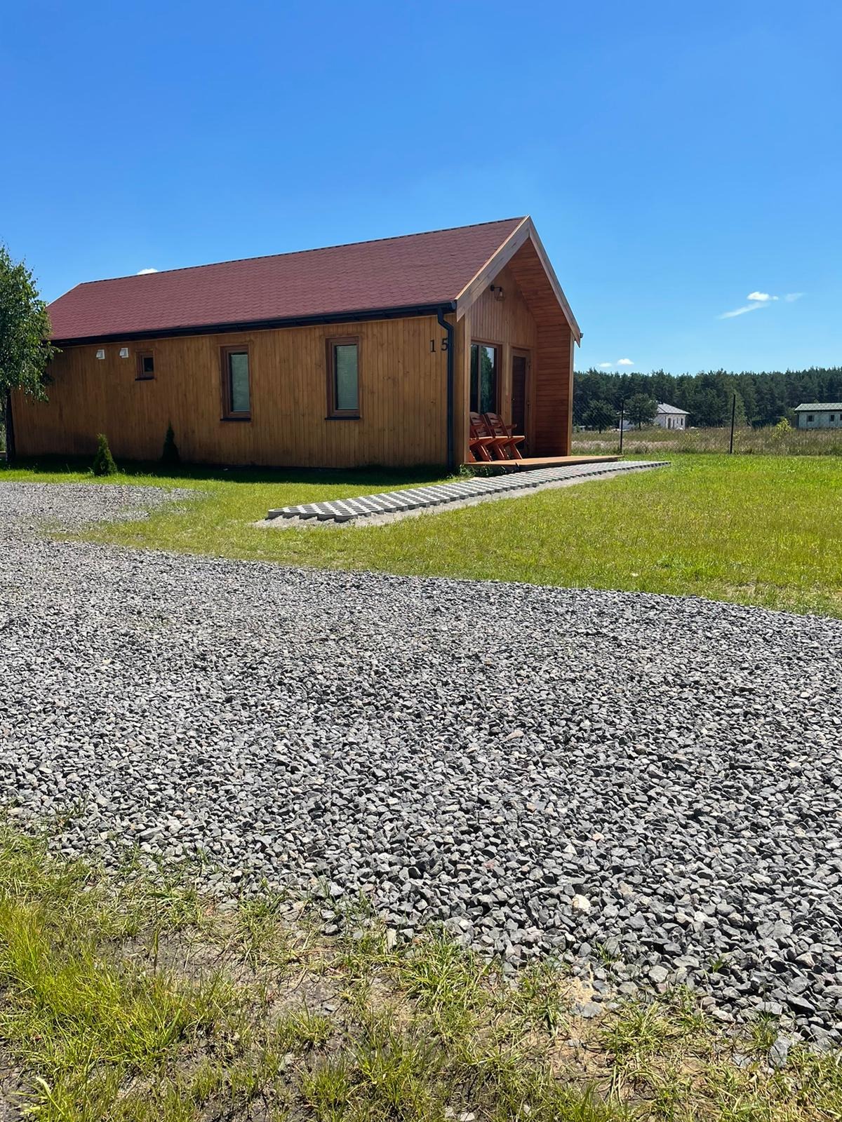 Nowoczesny, parterowy dom z drewna z czerwonym dachem, widok z zewnątrz na tle błękitnego nieba i zieleni, z żwirowym podjazdem i dwoma krzesłami na małym tarasie.