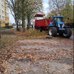 Niebieski traktor z czerwoną przyczepą stoi na placu z opadłymi liśćmi, obok leżą gałęzie po wycince, w tle blok mieszkalny.
