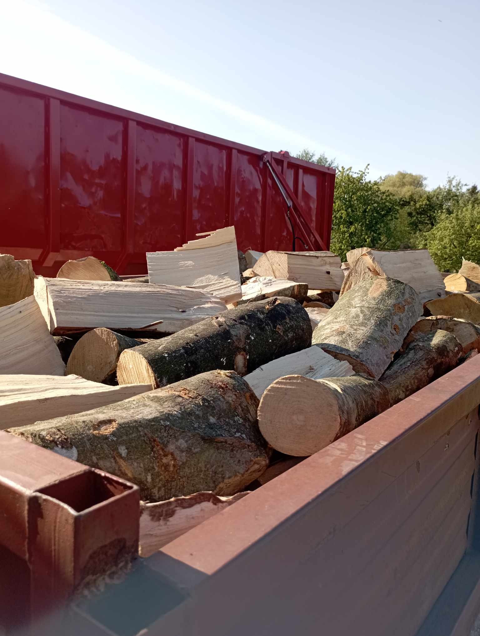 Ciężarówka z czerwonym kontenerem wypełniona pociętymi kawałkami drewna kominkowego o różnej grubości i fakturze kory, widoczne z bliska pod kątem.