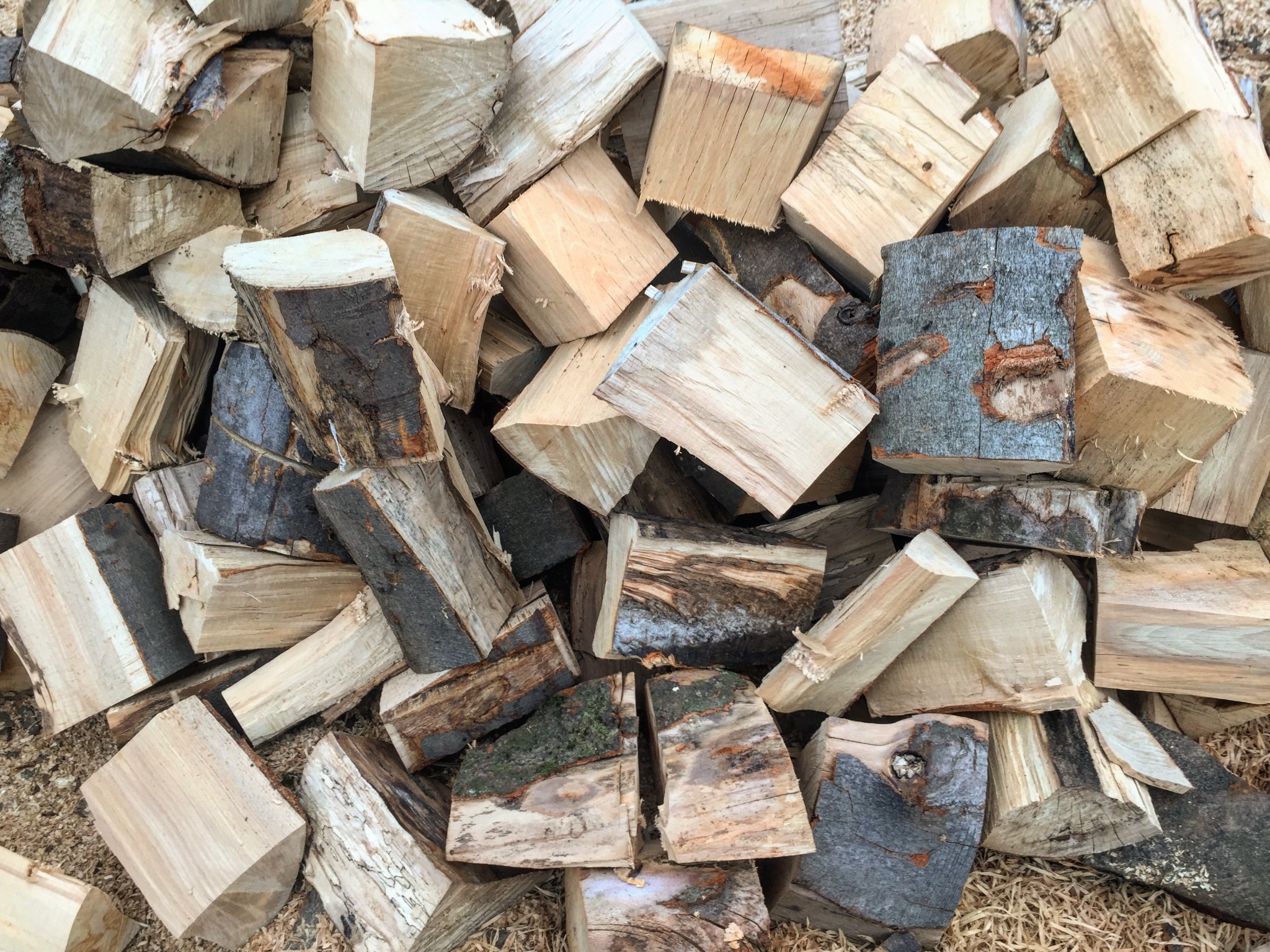 Stos pociętego drewna kominkowego o nieregularnych kształtach i różnej fakturze kory, ułożonego na podłożu z drobnymi wiórami.