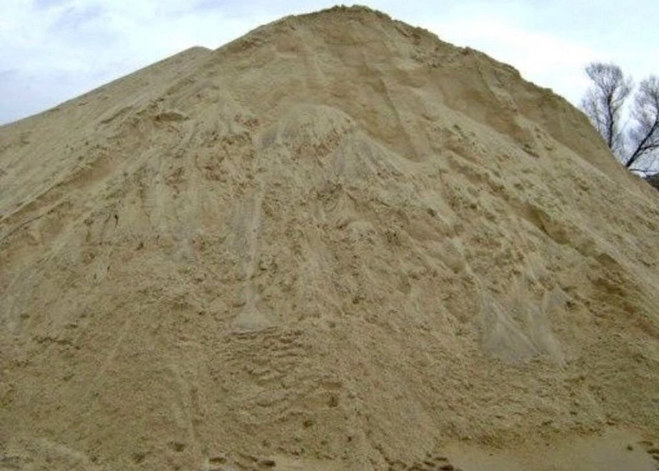 Duża pryzma piasku o drobnej strukturze, z widocznymi śladami po zsypywaniu, na tle pochmurnego nieba i fragmentu drzewa.