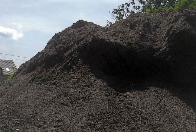 Duża pryzma ciemnej ziemi ogrodowej na tle nieba z widocznym dachem domu w oddali.