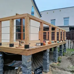 Drewniany taras w trakcie budowy, wsparty na kamiennych filarach, widoczne narzędzia, butelka wody na górnej krawędzi konstrukcji.