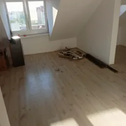 Poddasze w trakcie prac wykończeniowych: panele podłogowe ułożone, widoczne resztki materiałów i listwy przypodłogowe, okno dachowe z widokiem na budynki.